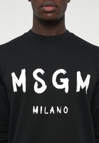 Schwarzer Rundhalsausschnitt-Sweatshirt mit fett gedruckten weißen "MSGM"-Buchstaben und kleinerem "MILANO"-Text darunter, getragen von einer Person, die bis zum Hals beschnitten ist.