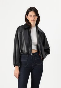 Giacca cropped in finta pelle nera con chiusura a zip, maniche oversize e collo a punta, abbinata a jeans a vita alta in denim scuro.