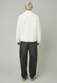 Personne avec les cheveux blonds courts portant une veste blanche, un jean noir décontracté et des chaussures blanches, se tenant de dos devant un fond uni.