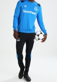 Blå sweatshirts med svarta detaljer, "SportPesa"-logotyp och klubbmärket; svarta byxor med blå sidopaneler; håller i en svart och vit fotboll.