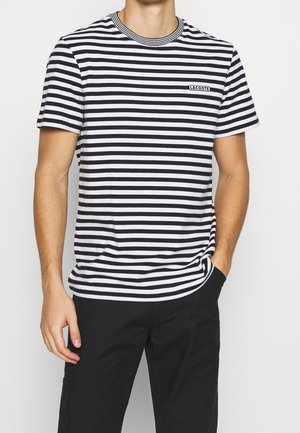 Mann trägt ein schwarz-weiß gestreiftes Kurzarmshirt mit "Lacoste"-Logo, kombiniert mit schwarzen Hosen, Hand in der Tasche.