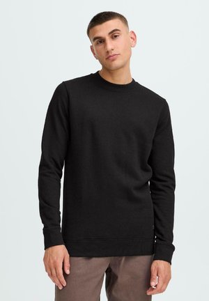 Zwarte sweatshirt met ronde hals van katoen. Lange mouwen, ribgebreide manchetten en zoom. Model staat met ontspannen handen langs de zij, gekleed in een broek in bruin.
