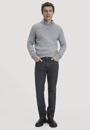 Grauer Strickpullover über einem hellblauen Hemd mit Kragen, kombiniert mit dunklen Denim-Jeans und schwarzen Knöchelstiefeln. Das Model steht mit den Händen in den Taschen.