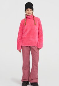 Fluffy roze coltrui met lange mouwen, gecombineerd met roze geruite broek en zwarte laarzen, tegen een witte achtergrond.