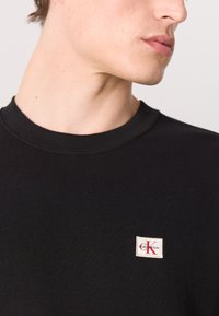 Zwarte katoen crewneck sweatshirt met een klein wit label met rode lettering op de borst. Soepele textuur en reguliere pasvorm.