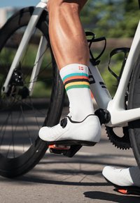 Zapatilla de ciclismo blanca con acentos negros y perforaciones, acompañada de un calcetín blanco con rayas horizontales multicolores, pierna sobre el pedal de la bicicleta.