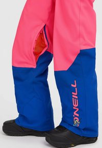 Heldere roze en blauwe sneeuwbroeken, met een mesh-voorgevoerde ritszak, en zwarte laarzen met een gestructureerde zool en logo-accenten op de broeken.