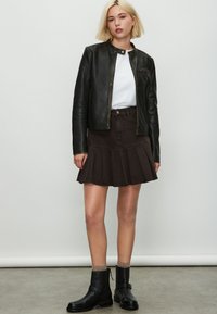 Giacca in pelle nera con collo rotondo, camicia bianca, gonna plissettata marrone e stivaletti neri con dettaglio fibbia. Colori e texture uniformi.