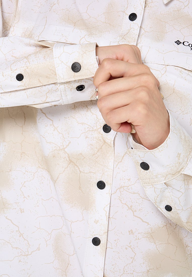 Weißes Hemd mit einem zerbrochenen beigen Muster, schwarzen Knöpfen und einem gekrempelten Ärmel. Verfügt über einen Kragen und gestickte Details auf dem Stoff.