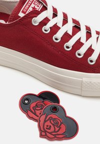 Röd kanvas sneakers med vita snören och metalldelar, med en vit gummisula. Två hjärtformade röda och svarta etiketter med rosdesign ligger i närheten.