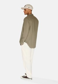 Camicia verde oliva con bottoni, dal taglio morbido, abbinata a pantaloni beige chiaro e sneaker nere, vista di lato.