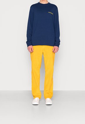 Marineblauw shirt met lange mouwen met gele tekst, gecombineerd met gele ribfluwelen broek en witte sneakers; gladde textuur en casual pasvorm.