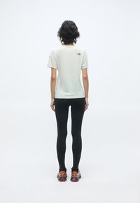 T-shirt beige clair à manches courtes avec un logo dans le dos, associé à des leggings noirs et des chaussures de sport avec des accents rouges.