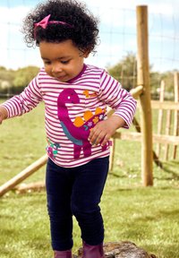 Gestreepte lange mouw shirt in roze en wit met een dinosaurus applicatie, gecombineerd met donkerg blauwe legging en paarse laarzen. Buitenomgeving.