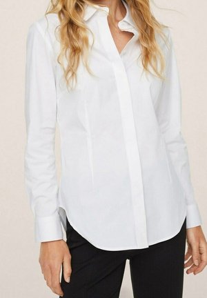 Kobieta ubrana w dopasowaną białą koszulę na guziki z długimi rękawami i czarne spodnie, stojąca na neutralnym tle.