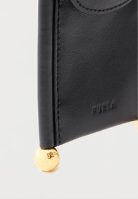 Melns ādas karšu turētājs ar iegravētu "Furla" logotipu, apaļiem zelta toņa metāla akcentiem stūros, gludu tekstūru un tīrām malām.