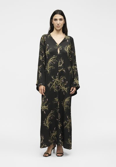 Γυναίκα με μακριά σκούρα μαλλιά που φοράει μαύρο μάξι φόρεμα με πράσινα και μπεζ φυτικά σχέδια και μαύρα σανδάλια με ψηλό τακούνι, στέκεται μπροστά σε λευκό φόντο.