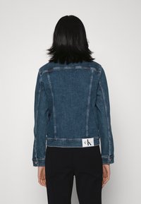 Modrá džínová bunda s přiléhavým střihem, viditelným šitím a nášivkou loga CK nad lemem. Klasický styl s dlouhými rukávy.