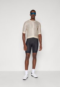 Mann in beigem Kurzarm-Radsporttrikot, schwarzen gepolsterten Radsportshorts, weißen Socken, weißen Radsportschuhen und blauen Sportsonnenbrillen, stehend.