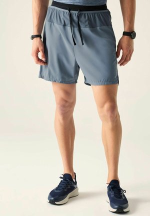 Mężczyzna ubrany w niebiesko-szare sportowe szorty z czarnym sznurkiem, granatowe buty do biegania i zegarek na nadgarstku, stoi na jasnym, jednolitym tle.