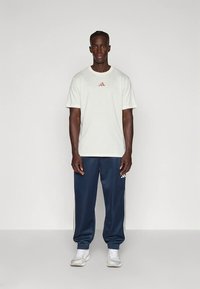 Hvit bomulls-T-skjorte med liten oransje Adidas-logo, kombinert med marineblå joggebukser med hvite sidestriper. Modellen står foran en nøytral bakgrunn.