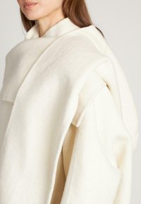 Cappotto oversized color crema in lana con una finitura testurizzata, dotato di un ampio colletto e di un tessuto drappeggiato che crea un effetto strati.