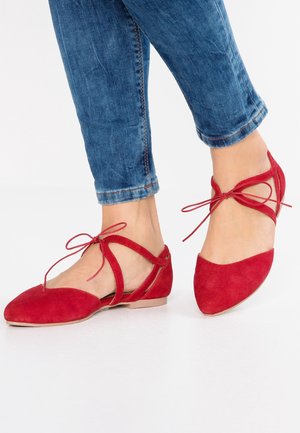 Czerwone zamszowe baleriny z szpiczastym noskiem, paskami krzyżującymi się na stopie oraz płaskim obcasem, zestawione z podwiniętymi niebieskimi dżinsami.