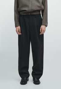 Schwarze, weit geschnittene Hosen mit glatter Textur, versehen mit einem Knopfverschluss und Gürtelschlaufen, kombiniert mit einem grauen Oberteil und schwarzen Schuhen.