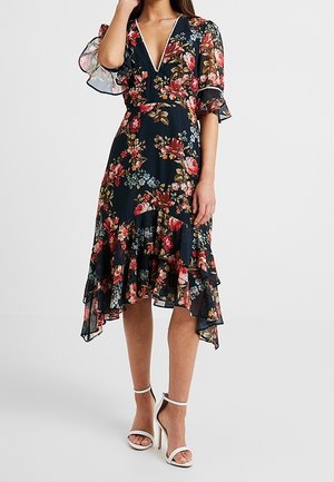 Kvinde iført navy blomster høj-lav kjole med gennemskinnelige flutterærmer og hvide ankelstroppesandaler med høje hæle, stående på ensfarvet baggrund.