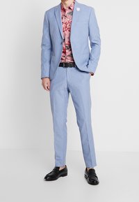 Lichtblauw pak met een smalle pasvorm, uitgevoerd in een gestructureerde stof, gecombineerd met een overhemd met bloemenpatroon in roze en zwart. Zwarte schoenen maken de look compleet.