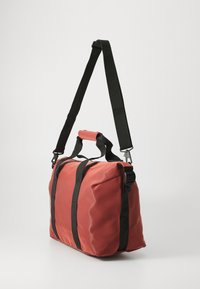 Sac de sport rouge avec des sangles noires, doté d'une poignée supérieure et d'une sangle d'épaule ajustable, sur fond blanc uni.