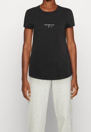 Osoba v černém tričku Calvin Klein Jeans a světle šedých teplácích stojí před jednoduchým světlejším pozadím.