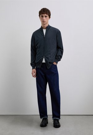 Chaqueta gris oscuro con cremallera y cuello alto, combinada con una camisa blanca, pantalones vaqueros azul oscuro y zapatos negros. Textura suave.