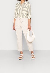 Světle béžové kalhoty s elastickým pasem, spárované s pruhovanou košilí v olivově zelené a bílé barvě. Bílá texturovaná kabelka a sandály na platformě.