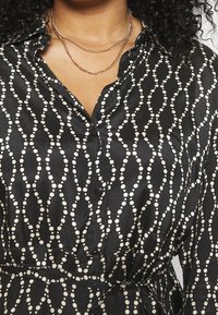 Czarna koszula w białe kropki, zapinana na guziki, z teksturowanym materiałem. Posiada kołnierzyk oraz pasek w talii dla lepszego dopasowania.