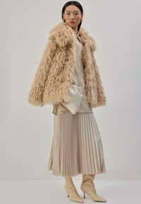 Beżowy płaszcz z futerka ekologicznego o puszystej fakturze i dużym kołnierzu, noszony na jasnobeżowej plisowanej spódnicy i kremowej obcisłej bluzce z kieszeniami.