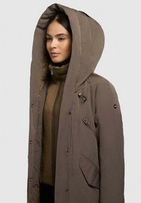 Chocoladebruine parka met een grote capuchon, opvallende ritssluitingen en drukknoopsluitingen. Bevat een gelaagde binnenjack voor extra warmte.