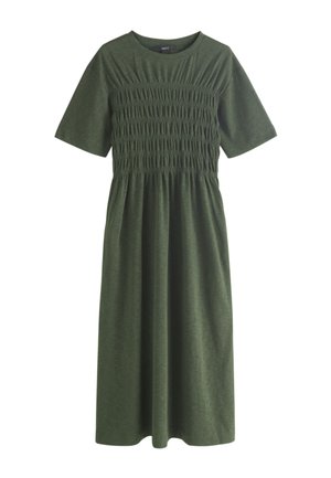 Ung kvinde går med en grå cropped jakke, mørkegrøn kjole i ankellængde og beige sandaler med sorte såler.