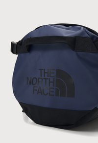 Round navy bag made of durable material with a black bottom. Features a prominent black logo and an adjustable strap.
