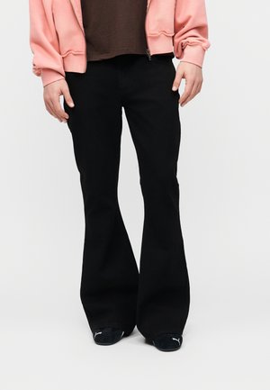 Άτομο που φοράει μαύρο παντελόνι με φούστα, μαύρα αθλητικά Puma, καφέ πουκάμισο και ροζ ζακέτα με φερμουάρ σε απλό λευκό φόντο.