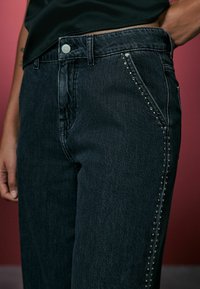 Czarne dżinsy o dopasowanej talii, z srebrnym guzikiem i ćwiekami wzdłuż kieszeni bocznych dla dodatkowego akcentu. Teksturowana tkanina.