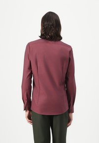 Chemise longue rouge bordeaux en tissu lisse, coupe structurée, motif subtil, avec des accents de boutons bleus aux poignets et au dos.