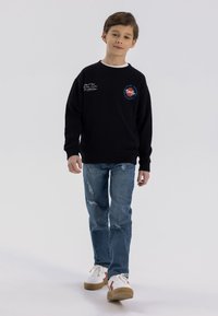 Sudadera negra con logotipo bordado, combinada con jeans destrozados de color azul claro y zapatillas blancas con detalles rojos.