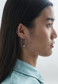 Krožna srebrna obročasta ušesna uhica z jasno kamenčki, ki vključuje majhen obesek; nosi se na levem ušesu ob ravnih temnih laseh.