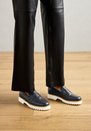 Pantalon en cuir noir longueur jambe, associé à des mocassins bleu marine avec des semelles épaisses blanches striées, sur un sol en bois.