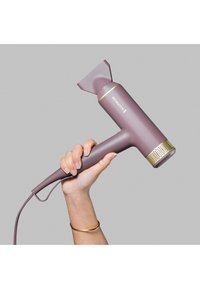 Peskegulv hårføner i ferskenfarge med en avsmalnende dyse, gullfarger og teksturert grep. Kabelen strekker seg fra håndtaket. Hånd som holder enheten.