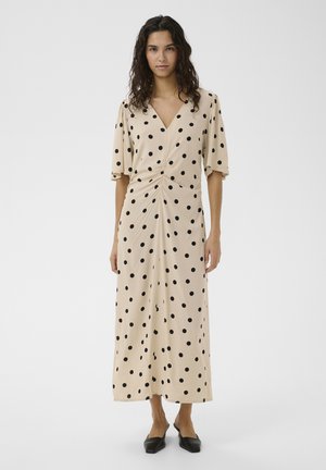 Kvinde stående iført en beige midi-kjole med sorte polkaprikker, rynket forside, korte ærmer, kombineret med sorte spidse sko.