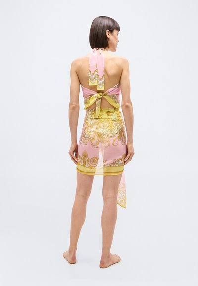 Γυναίκα σκυφτή, ξυπόλητη, με ροζ και κίτρινο φόρεμα με διάφανο μοτίβο, δέμενο στον λαιμό και στην πλάτη.