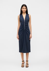 Γυναίκα που στέκεται με μπροστινή θέα, ντυμένη με αμάνικο navy midi φόρεμα με ρυτιδώσεις και φερμουάρ μπροστά, συνδυασμένο με μαύρα πέδιλα με λουράκια.