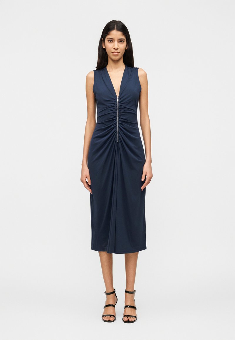 Γυναίκα που στέκεται με μπροστινή θέα, ντυμένη με αμάνικο navy midi φόρεμα με ρυτιδώσεις και φερμουάρ μπροστά, συνδυασμένο με μαύρα πέδιλα με λουράκια.